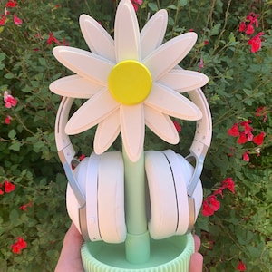 May include: A white daisy-shaped headphone stand with a green base and a yellow center. The stand is holding a pair of white headphones.