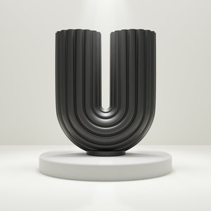 Peut inclure: Un vase décoratif noir en forme de U avec des rainures verticales, posé sur un socle circulaire blanc. Le vase a un design moderne et est présenté sur un fond neutre.