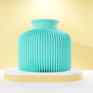 Pode incluir: Um vaso azul turquesa com um design canelado está em uma plataforma branca e amarela. O vaso está em uma superfície branca com um fundo amarelo.