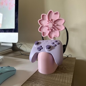 Pode incluir: Um suporte para controlador impresso em 3D rosa e roxo com um design de flor. O suporte segura um controlador roxo e fones de ouvido.