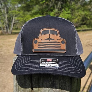 Puede incluir: Una gorra de camionero negra y gris con un parche de cuero marrón de una camioneta vintage. El parche tiene un diseño detallado de la parrilla delantera y los faros del camión. La gorra tiene una correa ajustable con el texto "Richardson 112 Original Trucker Adjustable" impreso en ella.