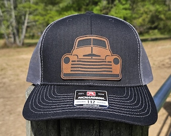 May include: A black and gray trucker hat with a brown leather patch of a vintage pickup truck. The patch has a detailed design of the truck's front grill and headlights. The hat has an adjustable strap with the text "Richardson 112 Original Trucker Adjustable" printed on it.
