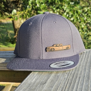 Könnte beinhalten: Graue Snapback-Kappe mit flachem Schirm. Auf der Kappe befindet sich ein Holz-Emblem eines Pick-up-Trucks auf der Vorderseite. Der Schirm ist in einem dunkleren Grauton gehalten, und die Kappe hat eine strukturierte Krone. Die Kappe liegt auf einer Holzoberfläche.