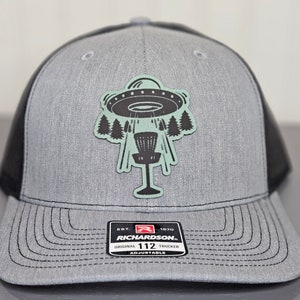 Może przedstawiać: Szara i czarna czapka trucker z naszywką przedstawiającą UFO przelatujące nad koszem do frisbee. Na czapce widnieje napis "Richardson" i "112".