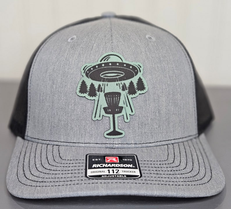 May include: A gray and black trucker hat with a patch of a UFO flying over a disc golf basket. The hat has the text "Richardson" and "112" on the front.