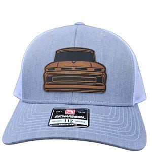 May include: A grey and white trucker hat with a brown leather patch depicting a vintage pickup truck. The patch features a black grille and headlights. The hat has the text "Richardson" and "112 Original Trucker" on the front.