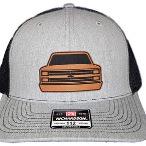 Può includere: Cappellino da camionista grigio con retro in rete nera e toppa in pelle marrone raffigurante la parte anteriore di un'auto. Il cappellino ha una visiera curva e un cinturino regolabile. La toppa mostra un contorno di auto nero con una griglia marrone. Il cappello ha il testo "RICHARDSON ORIGINAL 112 TRUCKER".