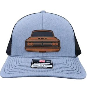 May include: A gray and black trucker hat with a brown leather patch of a GMC pickup truck on the front. The hat has a black mesh back and an adjustable strap with the Richardson 112 logo.