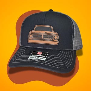 May include: A black and gray trucker hat with a brown leather patch featuring a vintage Ford truck silhouette. The hat has a black brim and a gray mesh back. The hat has a label that reads "Richardson. Est. 1970 Original 112 Trucker Adjustable".