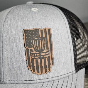 Könnte beinhalten: Eine graue und schwarze Trucker-Kappe mit einem braunen Leder-Patch, der einen Disc-Golf-Korb und ein amerikanisches Flaggen-Design zeigt.