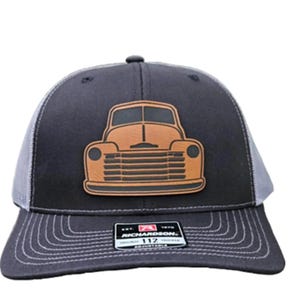 May include: A black and gray trucker hat with a brown leather patch of a vintage pickup truck. The hat has a mesh back and an adjustable strap with the text "Richardson 112" on it.