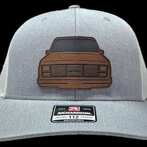 May include: A gray and white trucker hat with a brown leather patch of a vintage pickup truck. The patch has the text "GMC" on the front grill. The hat has an adjustable strap with the text "Richardson 112 Original Trucker Adjustable" on a red and white label.