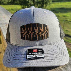 May include: Grey and black trucker hat with a brown leather patch reading "Dad". The hat features a mesh back and a curved peak. The Richardson 112 Trucker hat is adjustable.