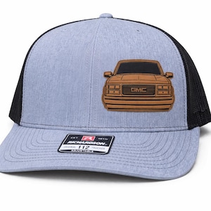 Puede incluir: Gorra de camionero azul claro y negro con un gráfico de camión GMC marrón en la parte delantera. La gorra tiene una visera curva y una correa ajustable. La etiqueta de la marca Richardson es visible.