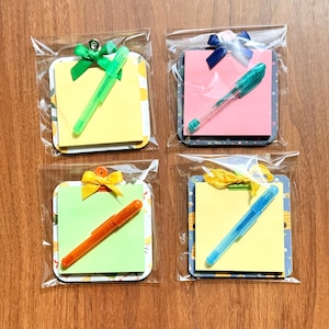 May include: Four square memo pads with colorful sticky notes, each featuring a matching plastic pen and a decorative ribbon bow attached to a metal clip. The sets are individually wrapped in clear plastic, showcasing yellow, pink, and light green paper.