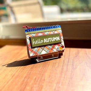 Könnte beinhalten: Ein kleiner Schreibtischkalender mit Spiralbindung und einem Karomuster in Orange, Grün und Weiß. Das Deckblatt zeigt ein dunkelgrünes rechteckiges Etikett mit dem weißen Text hello AUTUMN und einer kleinen Blumenverzierung.