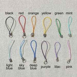 May include: A collection of colorful phone charms in black, red, orange, yellow, green, mint, light blue, sky blue, deep blue, purple, lilac, and pink. Each charm has a loop and a metal clasp, with color names labeled below.
