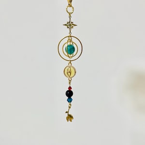 Puede incluir: Un charm dorado de tem&aacute;tica celestial con una estrella, c&iacute;rculos conc&eacute;ntricos y un orbe turquesa. Debajo, hay cuentas rojas, negras y azules, y un dise&ntilde;o de hoja dorada. El charm est&aacute; suspendido de un cierre dorado.