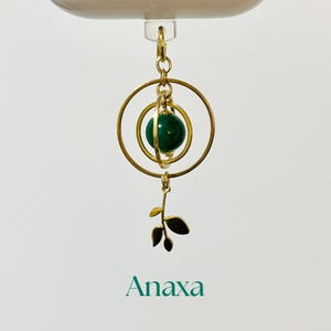 May include: A gold-colored pendant with a dark green sphere encased in a circular frame. A small leaf charm hangs below. The word "Anaxa" is printed in teal below the pendant.