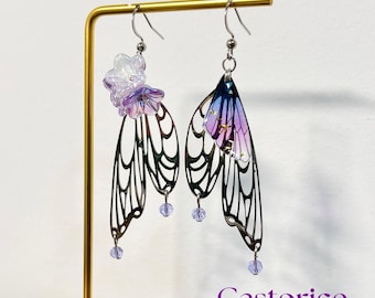 May include: A pair of dangle earrings with a fairy wing design. The earrings feature intricate metalwork in a dark bronze color, with translucent wings in shades of purple and blue. Each earring has a small flower and a dangling bead. The word "Castorice" is in the image.