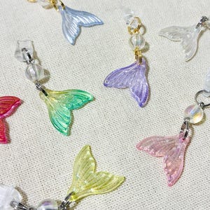 Peut inclure: Boucles d'oreilles avec des breloques en forme de queue de sirène aux couleurs vives : rose, violet, vert et jaune. Chaque breloque est fixée à une tige d'oreille transparente avec un anneau en métal argenté et une perle transparente. Finition translucide et irisée.
