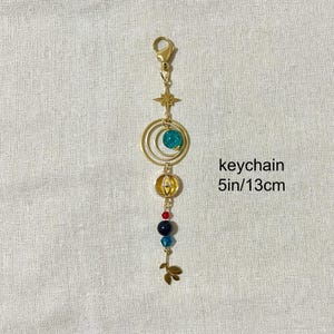 Puede incluir: Llavero dorado con una estrella, dos c&iacute;rculos conc&eacute;ntricos y una cuenta turquesa. El llavero tambi&eacute;n presenta un peque&ntilde;o dije redondo dorado y una serie de cuentas de colores. El texto "keychain 13cm" es visible.