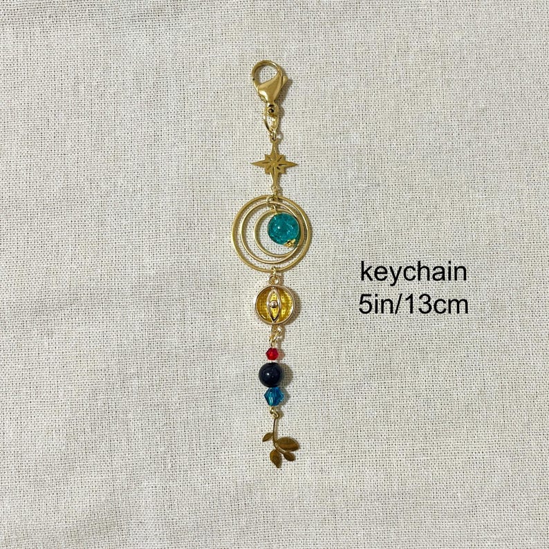 Puede incluir: Llavero dorado con una estrella, dos c&iacute;rculos conc&eacute;ntricos y una cuenta turquesa. El llavero tambi&eacute;n presenta un peque&ntilde;o dije redondo dorado y una serie de cuentas de colores. El texto "keychain 13cm" es visible.