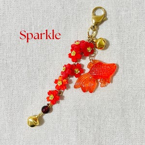 May include: A gold-toned keychain with a red floral design and an orange goldfish charm. The keychain also features a small gold bell and the word "Sparkle" in red text.