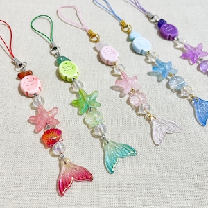May include: A collection of colorful mermaid-themed charms. Each charm features a fish, starfish, and mermaid tail design, strung with beads. The charms are in shades of pink, green, blue, and purple, with gold-colored accents and a cord for hanging.