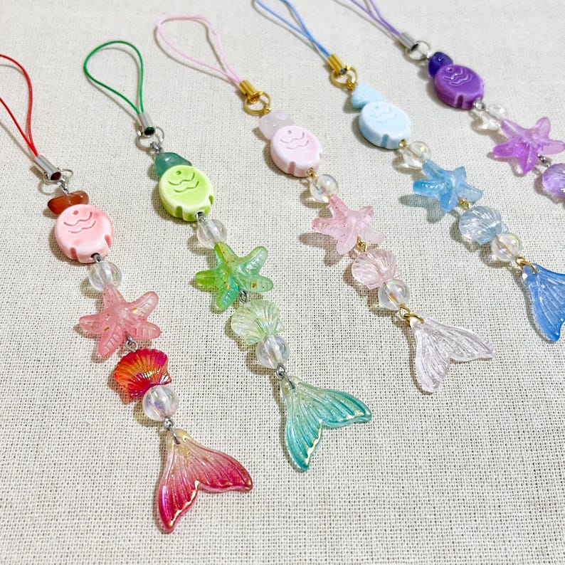 May include: A collection of colorful mermaid-themed charms. Each charm features a fish, starfish, and mermaid tail design, strung with beads. The charms are in shades of pink, green, blue, and purple, with gold-colored accents and a cord for hanging.