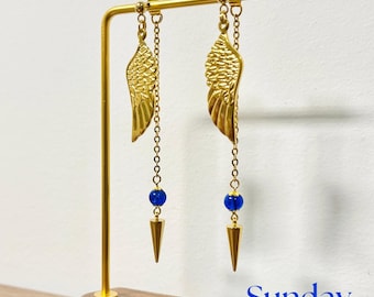 May include: Gold wing earrings with blue beads and gold cone-shaped tips hanging from a gold stand. The stand has a wooden base with the word "Sunday" written on it.