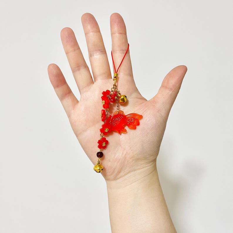 May include: A red and gold charm featuring a goldfish, flowers, and bells. The charm is suspended from a red cord and includes gold-colored accents. The goldfish has detailed fins and a flowing tail.