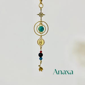 Puede incluir: Un dije dorado con una estrella, anillos y una esfera turquesa. Debajo hay cuentas rojas, negras y azules, y un dise&ntilde;o de hoja dorada. La palabra "Anaxa" est&aacute; impresa en verde.