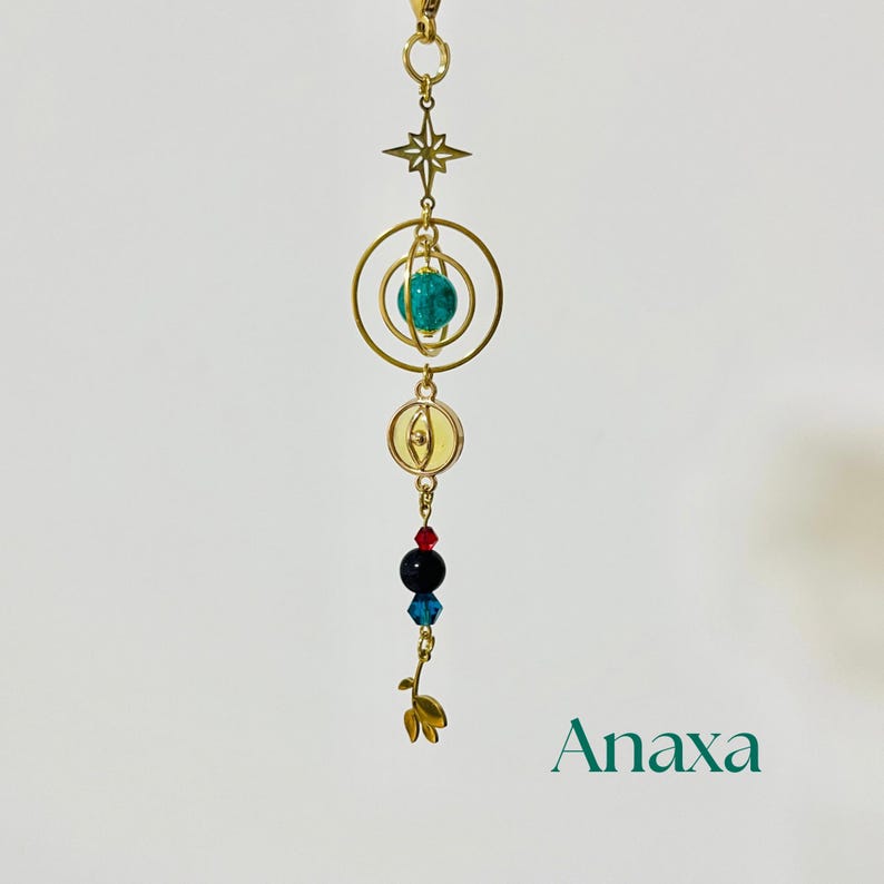 Puede incluir: Un dije dorado con una estrella, anillos y una esfera turquesa. Debajo hay cuentas rojas, negras y azules, y un dise&ntilde;o de hoja dorada. La palabra "Anaxa" est&aacute; impresa en verde.