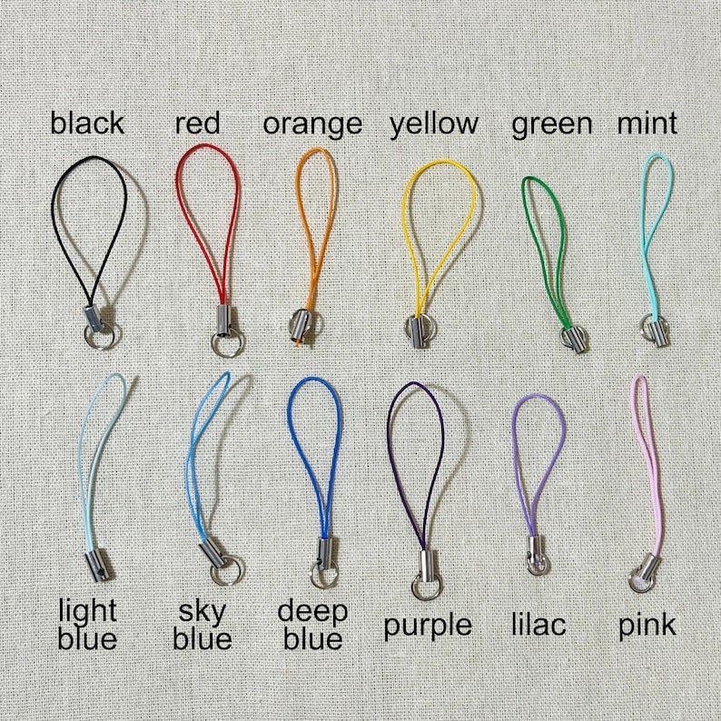 May include: A collection of colorful phone charms in black, red, orange, yellow, green, mint, light blue, sky blue, deep blue, purple, lilac, and pink. Each charm has a loop and a metal clasp, with color names labeled below.