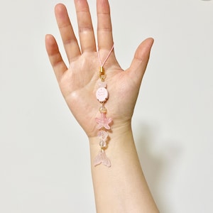 May include: A pink charm with a gold clasp and a pink cord. The charm features a pink cartoon character, a starfish, clear beads, and a mermaid tail. The charm is held in a hand.