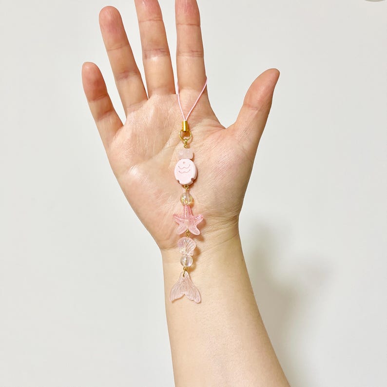May include: A pink charm with a gold clasp and a pink cord. The charm features a pink cartoon character, a starfish, clear beads, and a mermaid tail. The charm is held in a hand.