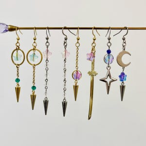 May include: A selection of dangle earrings with celestial and fantasy themes. Designs include stars, moons, swords, and geometric shapes. The earrings feature gold, silver, and black metal with colourful beads and crystal accents. Each earring has a unique design and a pointed bottom.