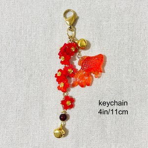 May include: A gold-colored keychain with a red goldfish charm, red flower accents, and a gold bell. The keychain is approximately 4 inches (10 cm) long.
