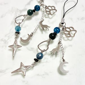 May include: Two silver-toned phone charms featuring blue and black beads, a heart-shaped link, a crescent moon charm, a wolf silhouette charm, a paw print charm, and a four-pointed star charm. The charms are attached to a black cord loop.