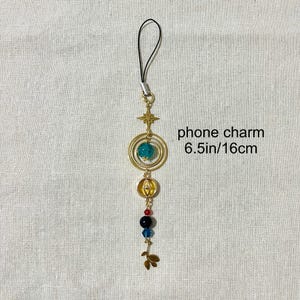 Puede incluir: Un charm de tel&eacute;fono dorado con una correa negra. El charm presenta una estrella, c&iacute;rculos conc&eacute;ntricos con una cuenta azul, un elemento en forma de calabaza y cuentas de colores. El texto "phone charm 6.5in/16cm" es visible.