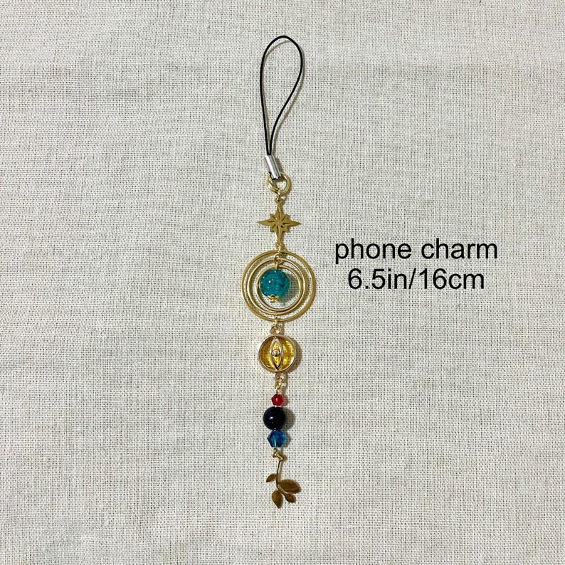 Puede incluir: Un charm de tel&eacute;fono dorado con una correa negra. El charm presenta una estrella, c&iacute;rculos conc&eacute;ntricos con una cuenta azul, un elemento en forma de calabaza y cuentas de colores. El texto "phone charm 6.5in/16cm" es visible.
