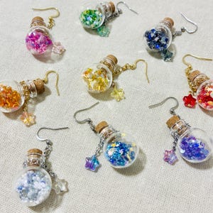 May include: A collection of handmade earrings featuring small glass bottles filled with colorful glitter. Each bottle has a cork stopper, a chain, and a star-shaped charm. The earrings come in various colors, including pink, green, blue, and gold.