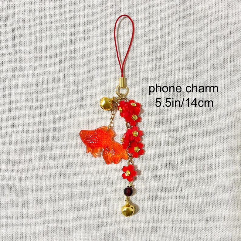 May include: A phone charm featuring a vibrant orange goldfish, red flowers, and gold accents. The charm includes a small bell and a red loop for attachment. The text "phone charm 5.5in/14cm" is visible.