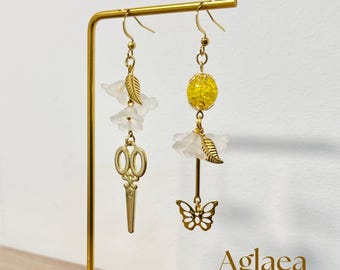 May include: A pair of gold-colored dangle earrings. One earring features white flower accents and a gold scissor charm. The other earring has a yellow bead, white flower accents, a gold leaf, and a gold butterfly charm. The word "Aglaea" is in the bottom right corner.