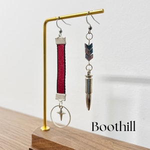 May include: A pair of earrings on a display stand. The left earring features a red ribbon with a silver star charm. The right earring features a silver bullet charm with a silver arrow charm above it. The text "Boothill" is visible in the bottom right corner of the image.
