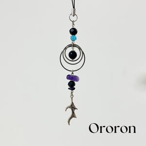 May include: A silver-toned charm with a black cord. The charm features black and blue beads, three silver rings, and a silver bat-wing design. The word "Ororon" is printed in black at the bottom of the image.