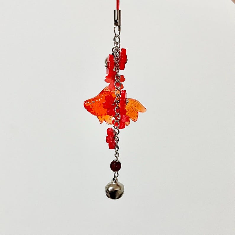 May include: A red and orange goldfish charm with a silver chain, small red flower accents, a dark red bead, and a silver bell. The charm is suspended from a red cord, creating a decorative accessory.