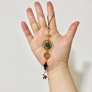 Puede incluir: Un dije dorado con una estrella, c&iacute;rculos conc&eacute;ntricos y cuentas de colores. El dije presenta una cuenta verde, una cuenta naranja y varias cuentas rojas, negras y azules m&aacute;s peque&ntilde;as. Una cuerda negra est&aacute; unida en la parte superior.