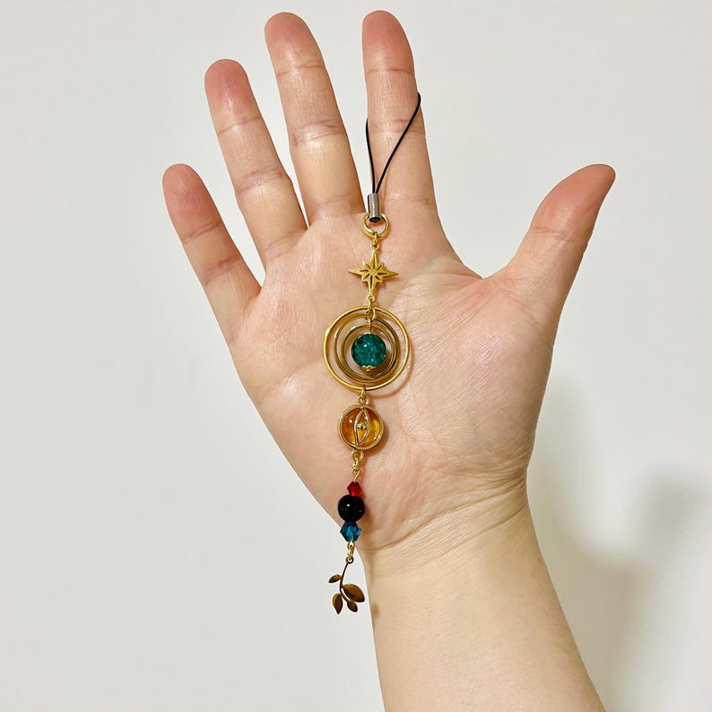 Puede incluir: Un dije dorado con una estrella, c&iacute;rculos conc&eacute;ntricos y cuentas de colores. El dije presenta una cuenta verde, una cuenta naranja y varias cuentas rojas, negras y azules m&aacute;s peque&ntilde;as. Una cuerda negra est&aacute; unida en la parte superior.